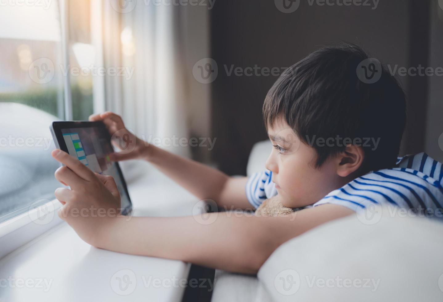 Cinematic portrait young boy playing game on tablet sitting on sofa with light shining from window, Kid playing games on internet, Child doing homework online at home photo