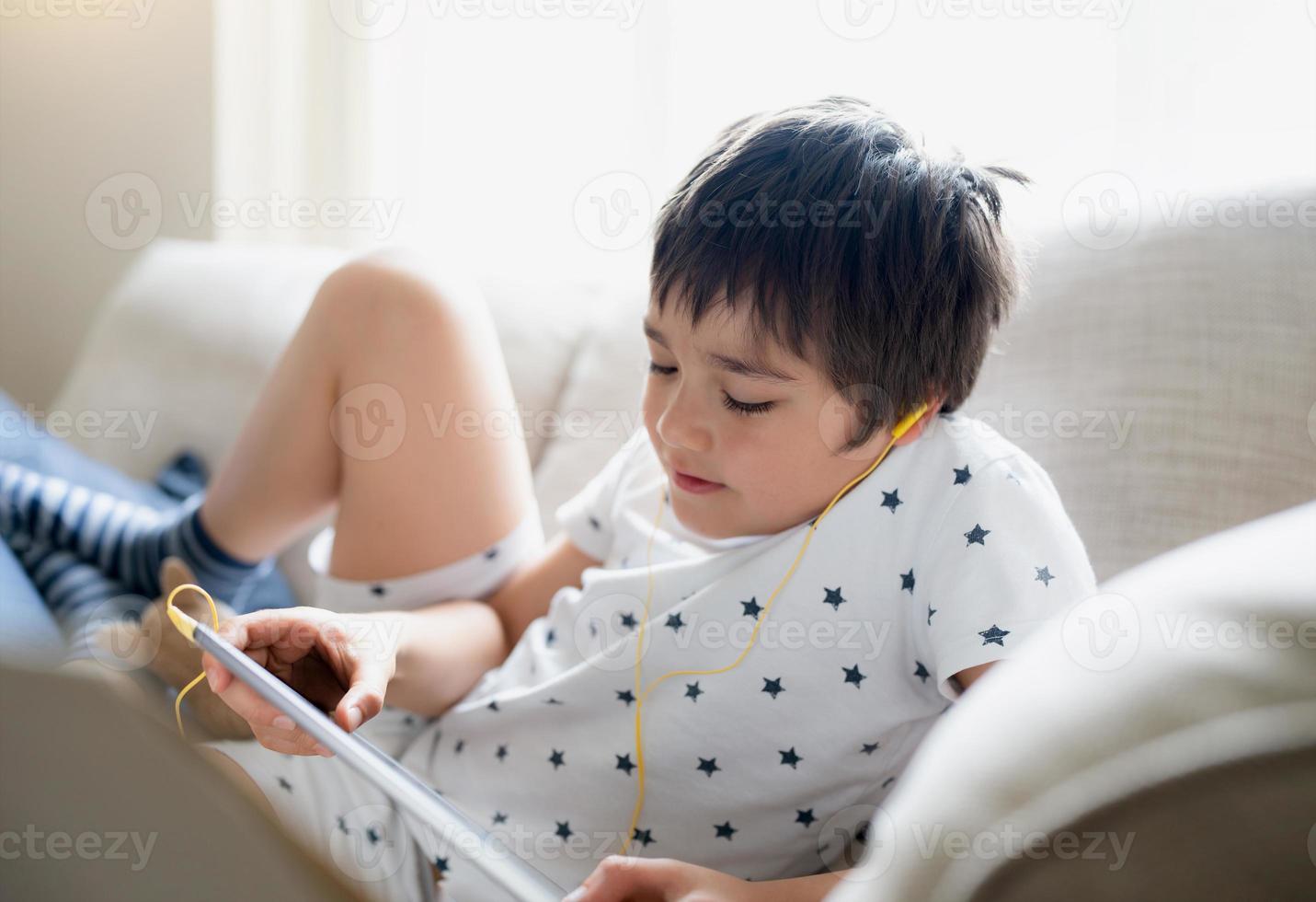 High key portrait kid wearing earphones listening to music, playing game on tablet,Boy sitting on sofa with light shining from window, Kid playing games on internet,Child doing homework online at home photo