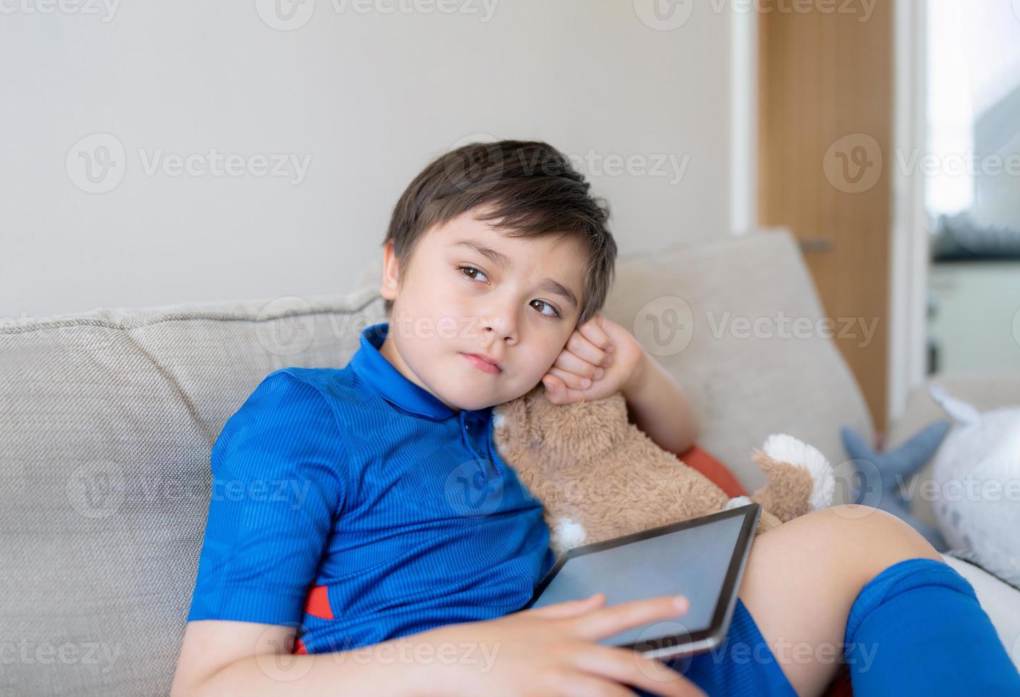 Young boy holding tablet mockup and looking deep in thought, Child playing game online on digital pad, School kid with sportswear sitting on sofa relaxing after play soccer or football with friends photo