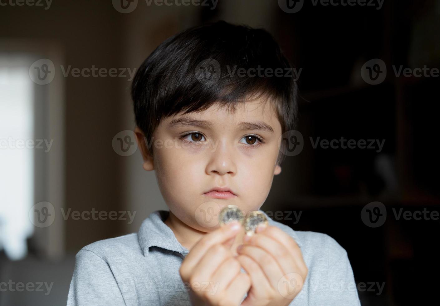 Portrait School Kid looking out deep in though, Selective focus Child boy holding one pound coin looking out with sad face, Bored Child standing alone in living room, Children learning to save money photo