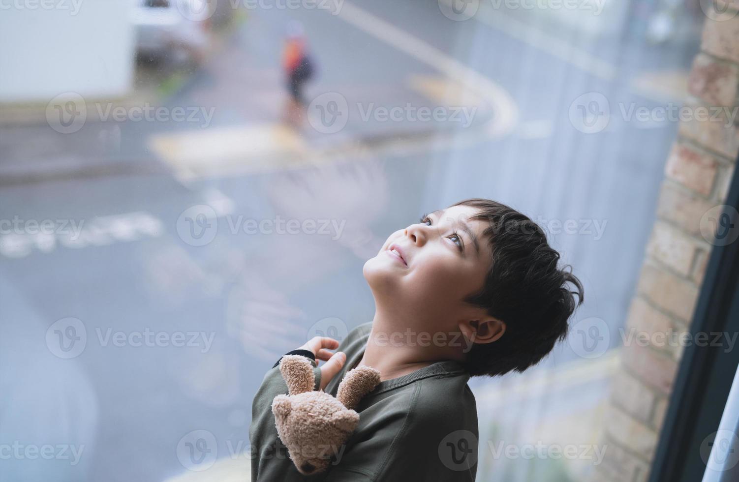 Young kid standing next to window looking up to sky with smiling face, Child looking through window glass with wondering face. Portrait boy relaxing in room apartment in city photo