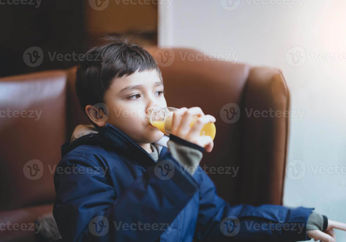 Healthy Kid boy drinking fresh orange juice in the cafe with bright light shining from window, Child sitting on sofa in coffee shop looking out with thinking face while drinking juice after snack. photo