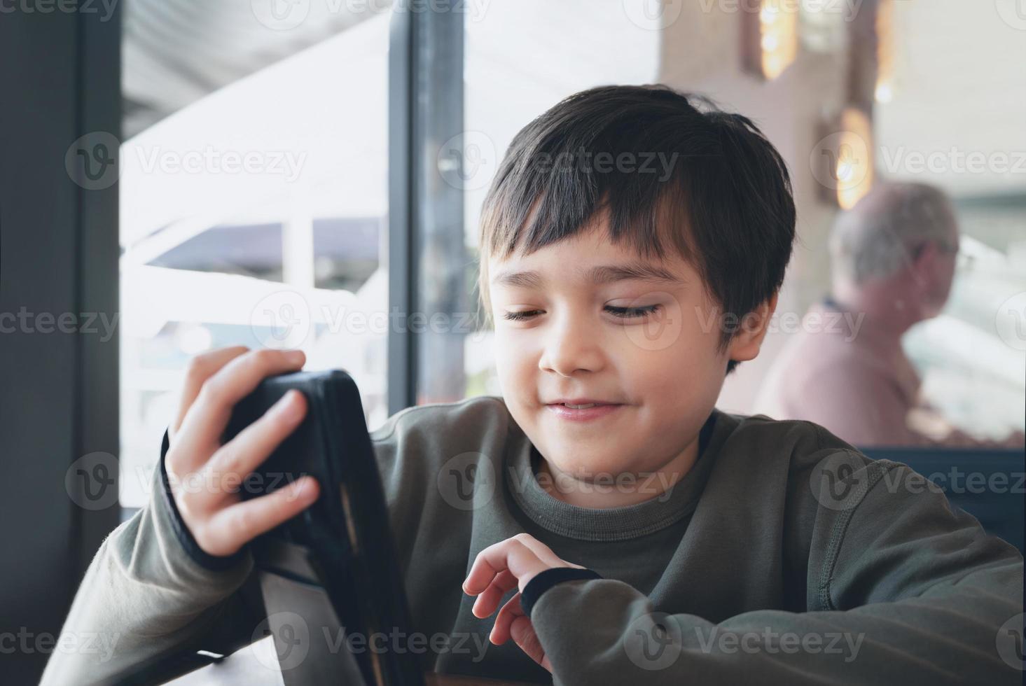 Cenimatic portrait young boy playing game on mobile while waiting for food, Kid sitting in coffee shop sending text to friends, Child playing game online on phone. photo