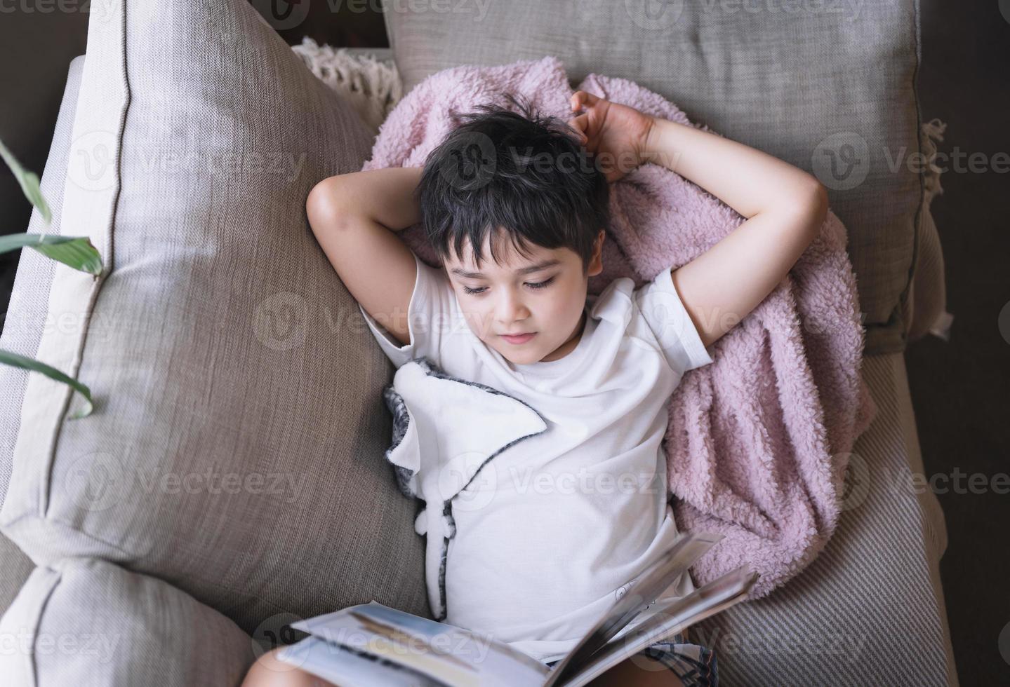 Happy School kid lying on sofa reading a book with morning light shining from window. Top view Child boy reading story relaxing at home on weekend, Positive children concept photo