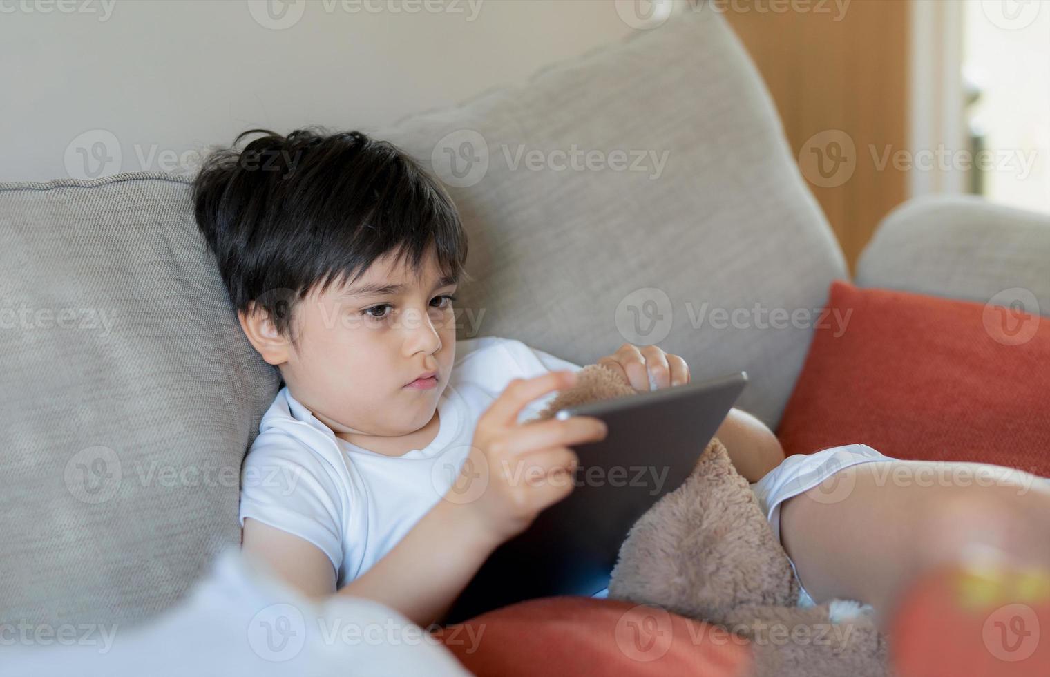 Young boy holding tablet looking out deep in thought, Unhappy kid with sad face sitting alone on sofa, A lonely child playing with soft toy and playing game on digital pad at home. photo