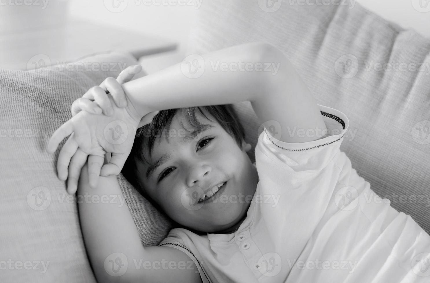 Portrait black and white image of happy young boy looking at camera with smiling face, Positive child lying on sofa relaxing at home. Head shot mono tone of smile kid photo