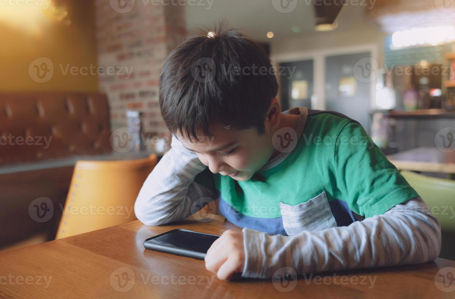 Cinematic portrait young boy playing game on mobile while waiting for food, Kid sitting in coffee shop sending text to friends, Child playing game online on phone.Children with technology concept photo
