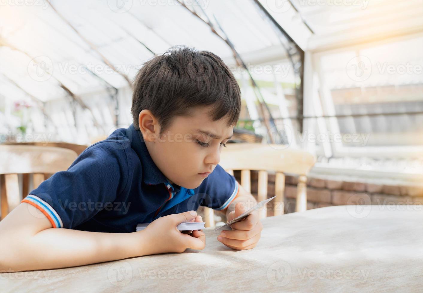Young boy playing card game in the cafe while waiting for food, Kid with curious face looking at paper card on his hands,  Family playing board game indoor on sunny day spring or Summer. photo