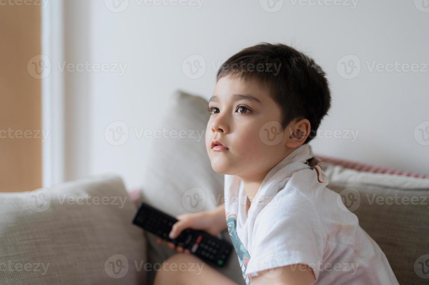 Portrait young boy holding remote control and looking up with curious face, Kid sitting on sofa watching cartoon on TV, Child lying down on couch relaxing in living room after back from school. photo