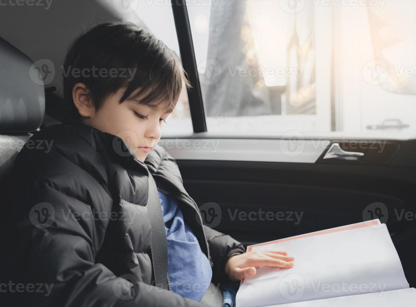 Kid siting on car seat and reading a book, Young boy sitting in  safety seat, Portrait of child entertaining himserf on a road trip. Concept of safety taveling by car with children photo