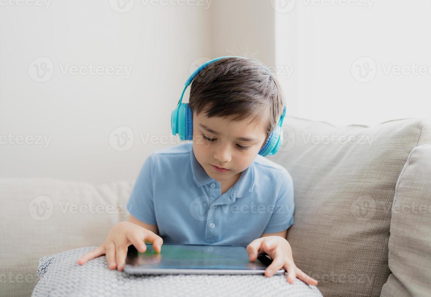 niño feliz usando auriculares para jugar en Internet con amigos, niño sentado al lado de la ventana leyendo o viendo dibujos animados en la tableta, niño de la escuela haciendo la tarea en línea en casa. foto