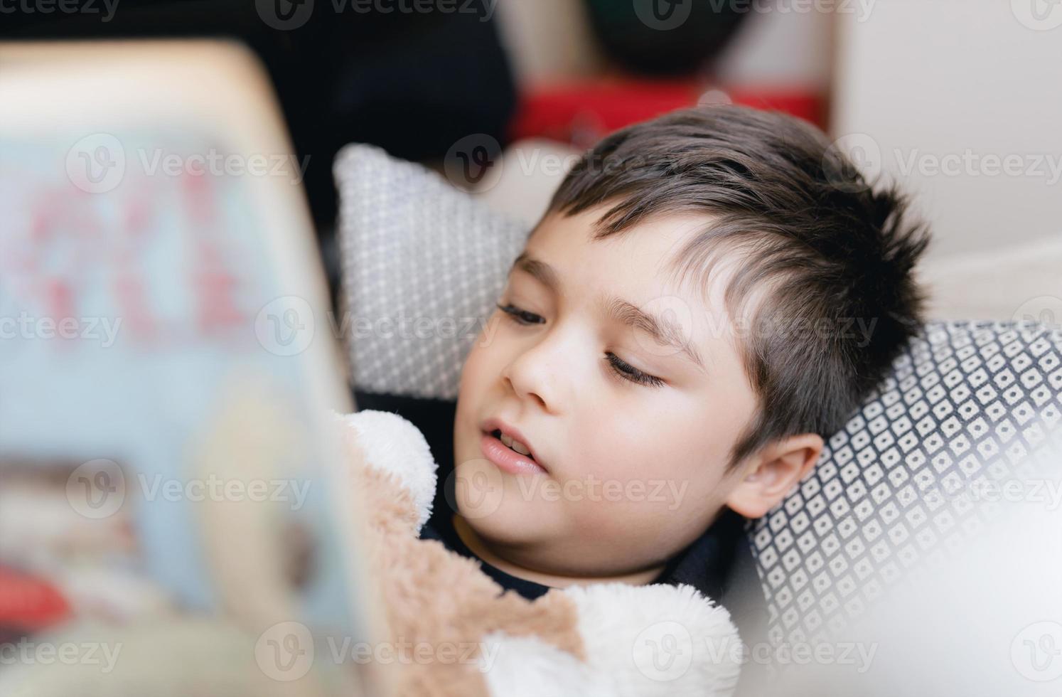 niño de la escuela de cara cerrada leyendo un libro para la tarea, tiro en la cabeza niño acostado en el sofá disfruta leyendo la historia en la sala de estar, niño relajándose en casa en la primavera soleada, educación en el hogar, concepto de educación foto