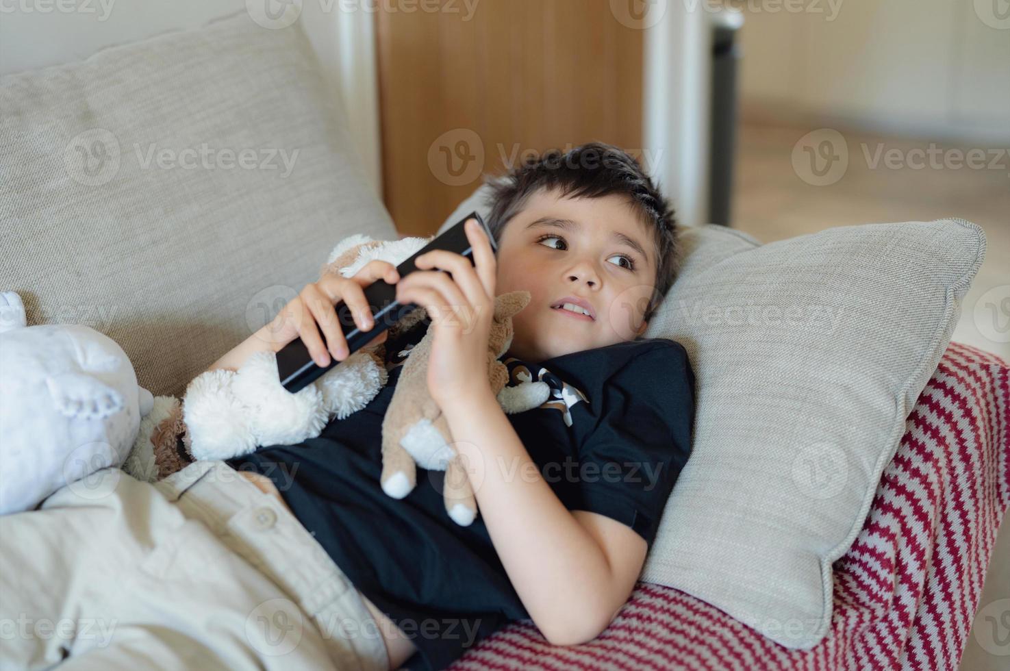 Happy Young boy holding remote control and looking up with smiling face, Cute kid lying on sofa enjoy watching TV program, Child relaxing at home on weekend photo