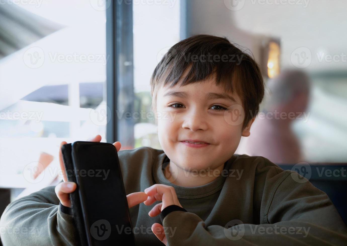 Cinematic portrait young boy playing game on mobile while waiting for food, Kid sitting in coffee shop sending text to friends, Child playing game online on phone. photo