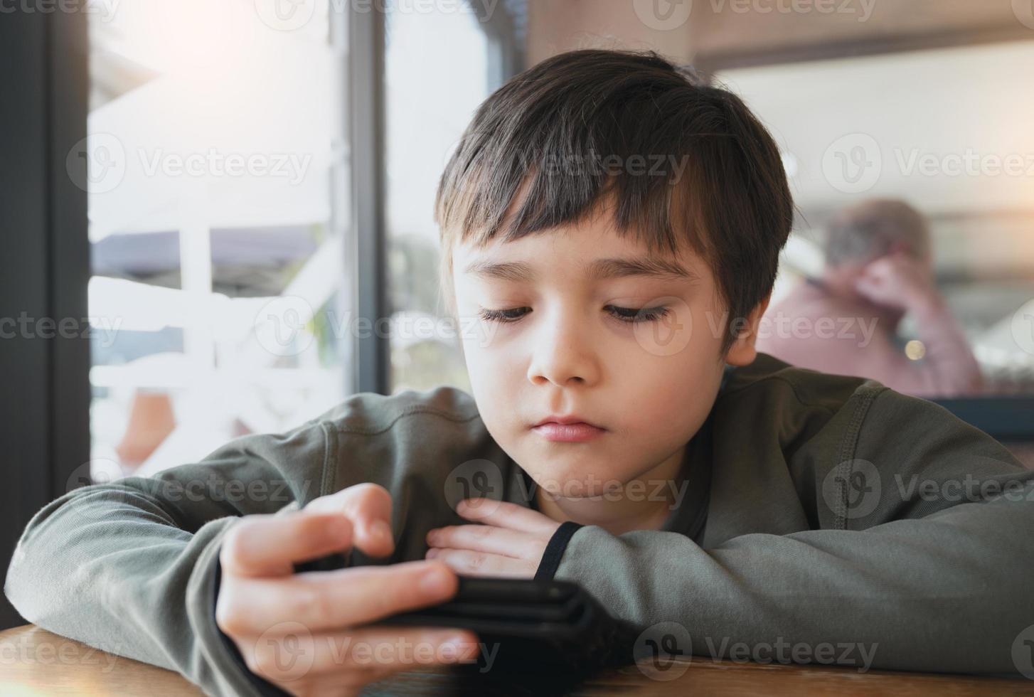 Cinematic portrait young boy playing game on mobile while waiting for food, Kid sitting in coffee shop sending text to friends, Child playing game online on phone. photo