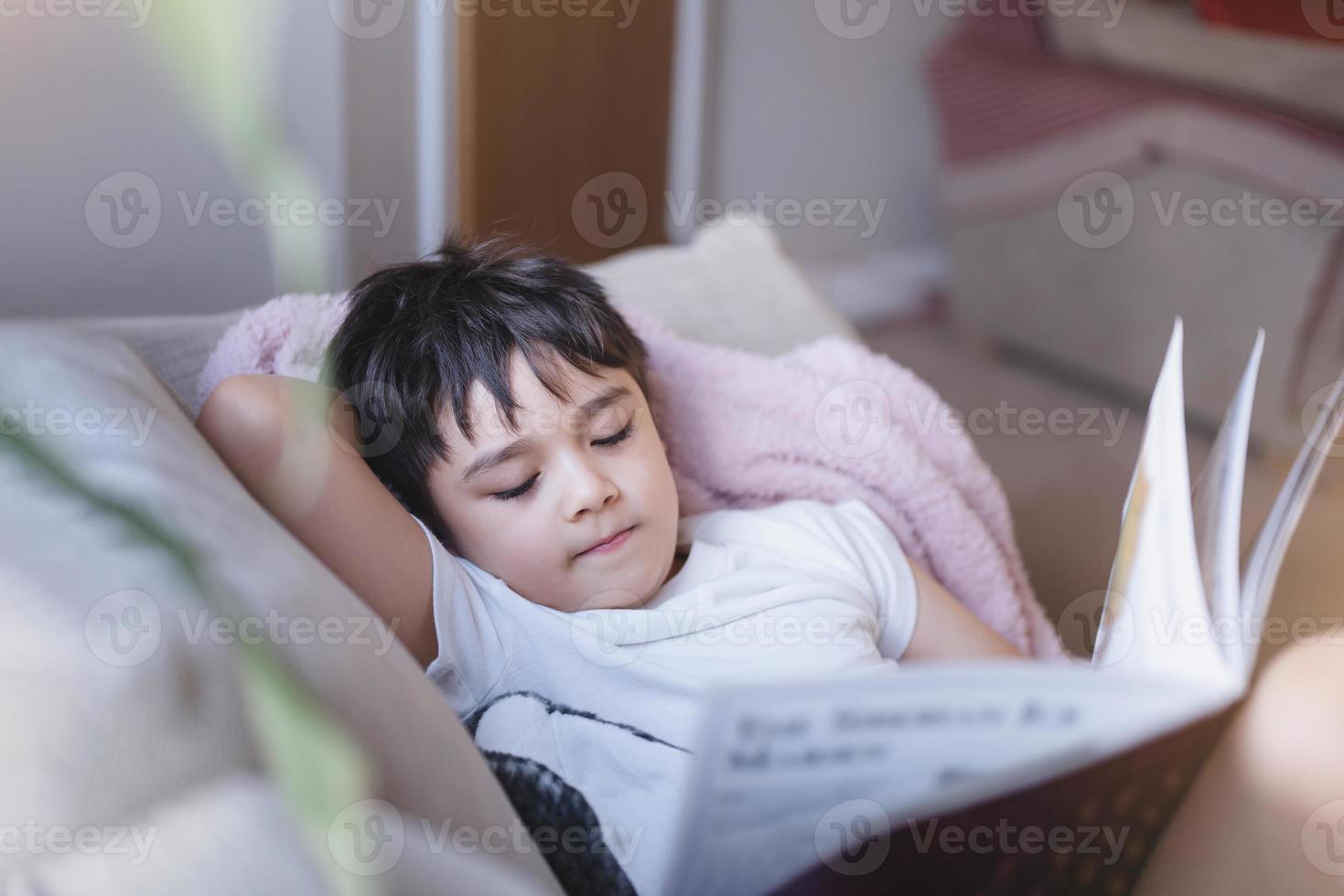 niño de la escuela feliz acostado en el sofá leyendo un libro con la luz de la mañana brillando desde la ventana. niño niño leyendo historia relajándose en casa el fin de semana, concepto de niños positivos foto