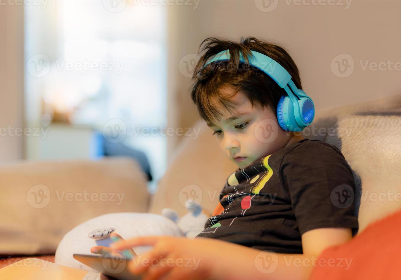 Happy young boy wearing headphone for playing game online on internet with friends, Kid sitting on sofa reading or watching cartoon on tablet Child relaxing at home in the morning on weekend photo