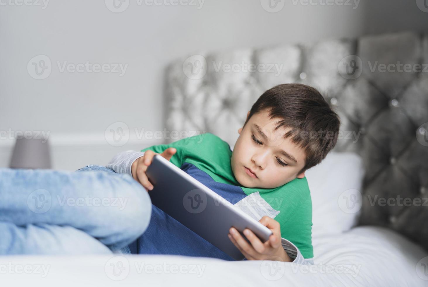 niño usando una tableta jugando en Internet con amigos, niño educando en el hogar haciendo la tarea en línea con una libreta digital en casa, niño acostado en la cama relajándose, viendo dibujos animados o hablando en línea con un amigo foto