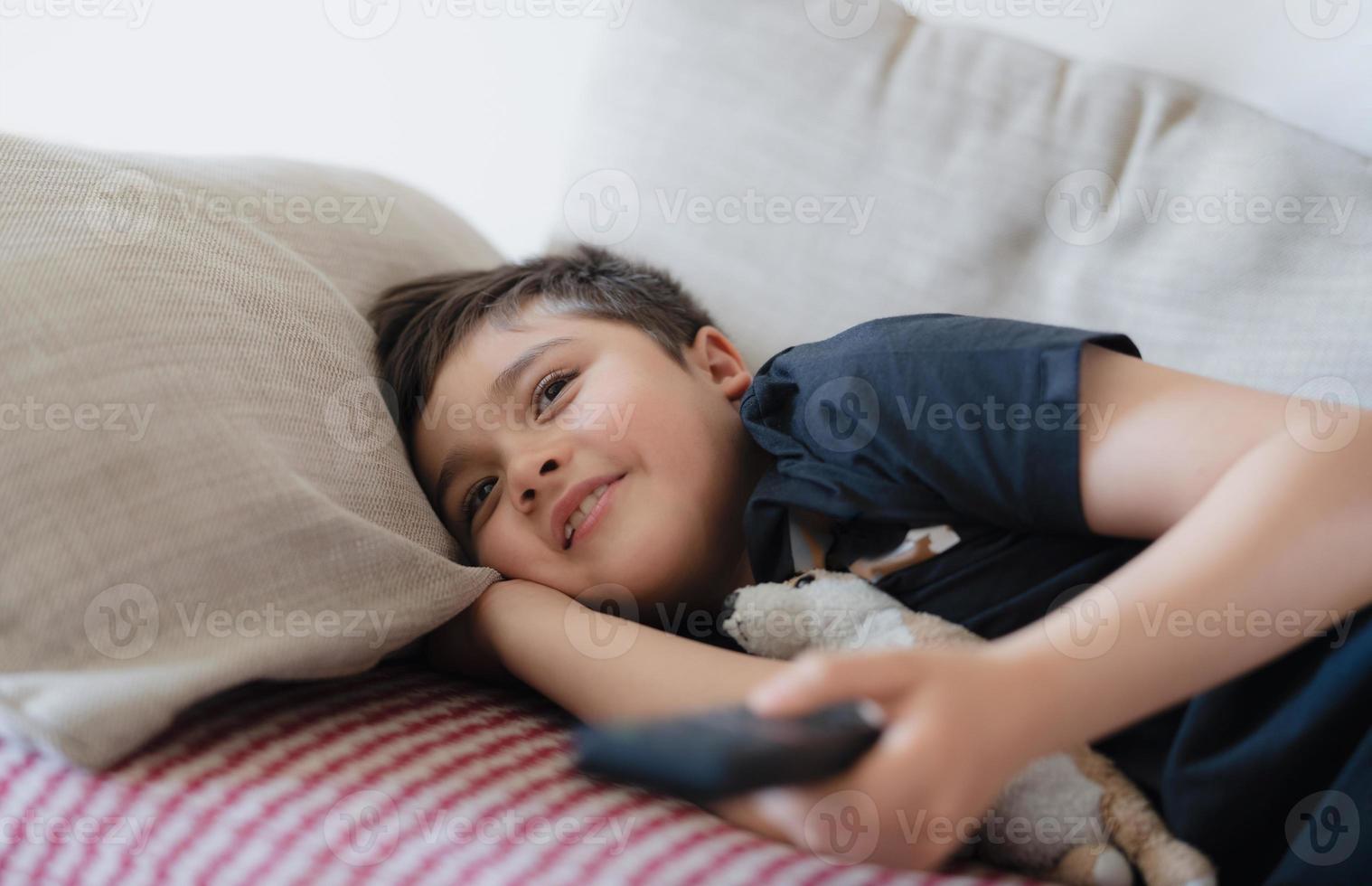 Happy Young boy holding remote control and looking up with smiling face, Cute kid lying on sofa enjoy watching TV program, Child relaxing at home on weekend photo