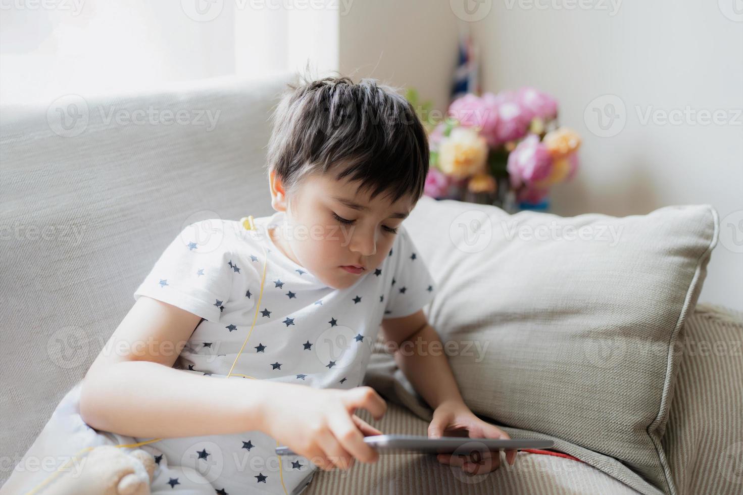 High key portrait kid playing game on tablet sitting on sofa with light shining from window, Young boy  playing games on internet, Cinematic portrait Child doing homework online at home photo