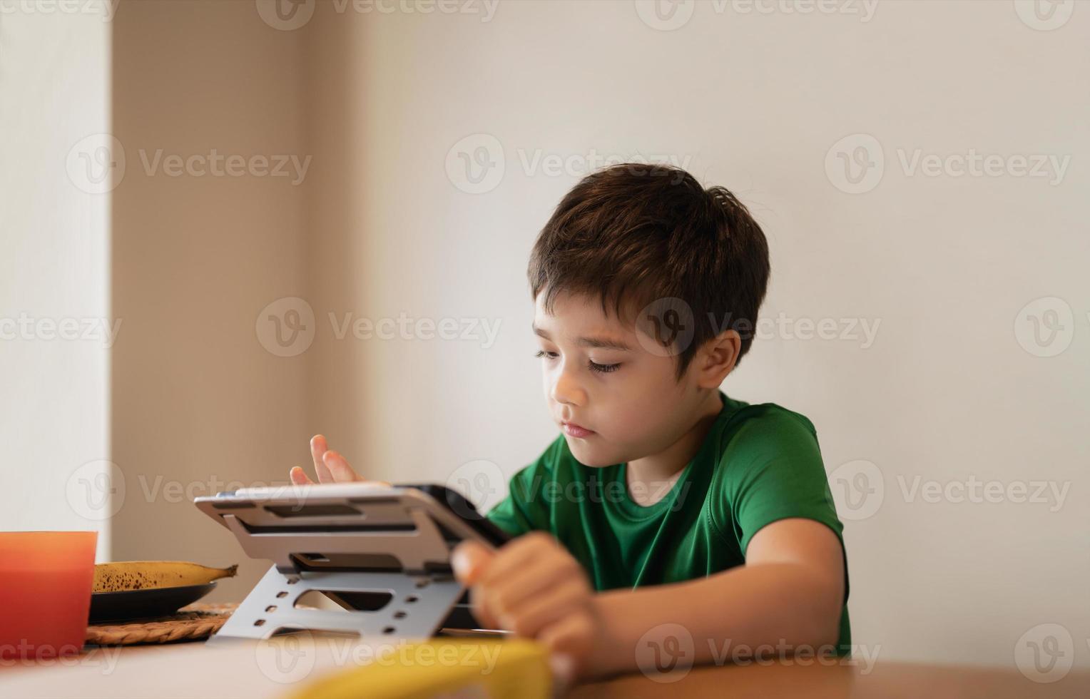 Kid playing game online with friends on tablet, Cinematic portrait young boy video call with friends while having breakfast,Child watching cartoon on internet, School kid doing homework on digital pad photo