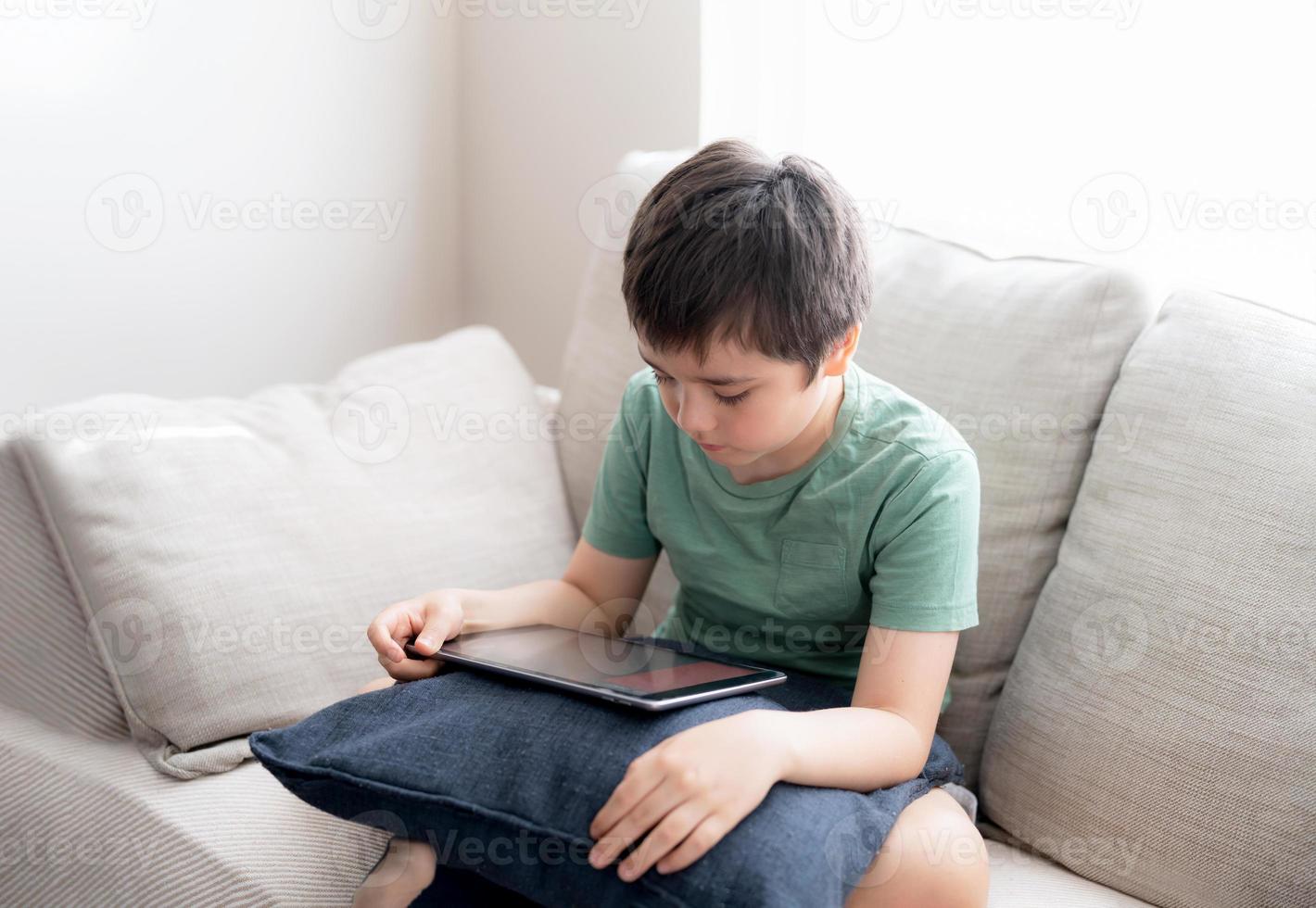 High key portrait young boy playing game on tablet sitting on sofa with light shining from window, Kid playing games on internet, Cinematic portrait Child doing homework online at home photo