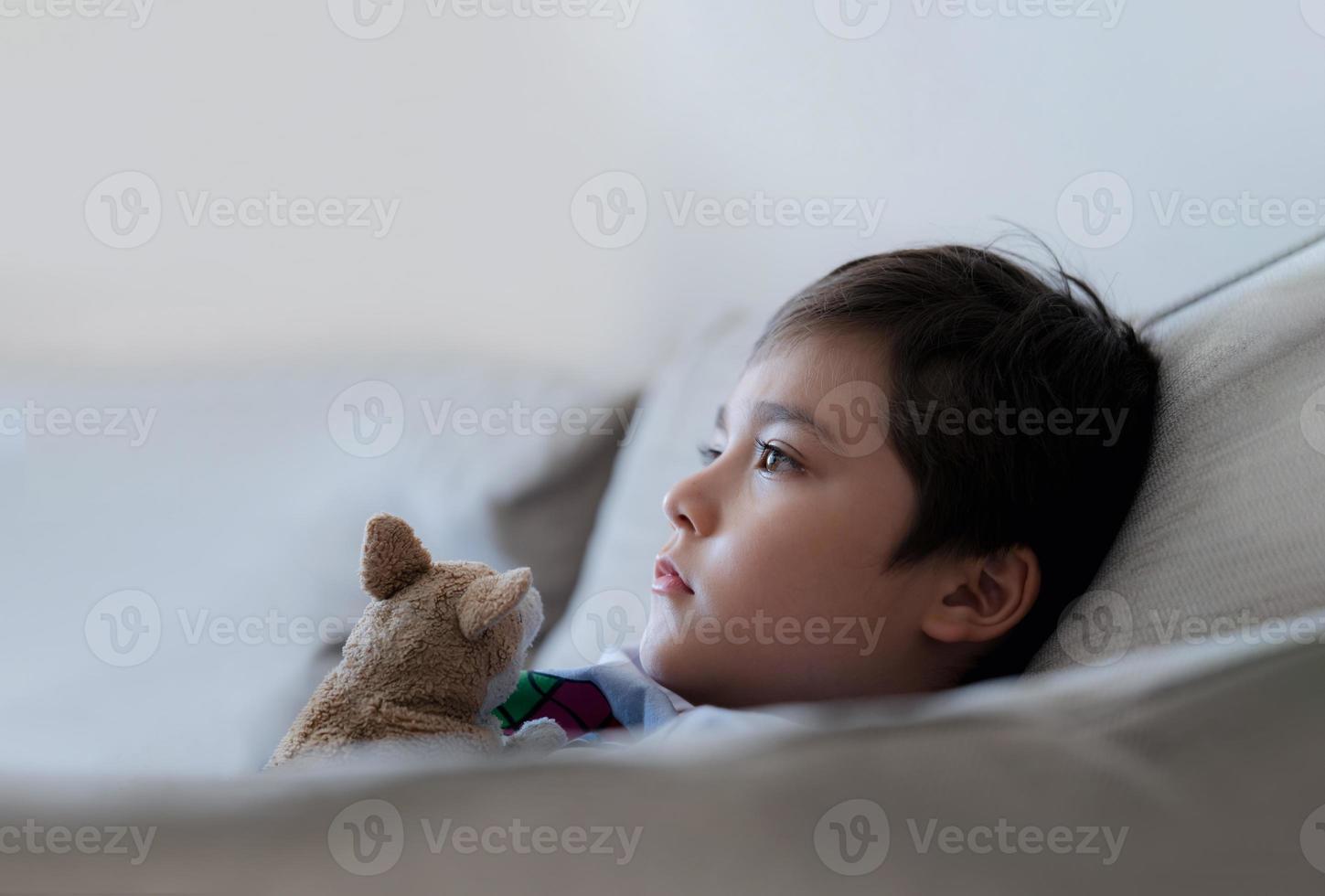 cerrar la cara niño sentado en el sofá con juguete de perro viendo la televisión, niño sentado en el sofá mirando profundamente en el pensamiento. niño positivo relajándose en casa el fin de semana foto