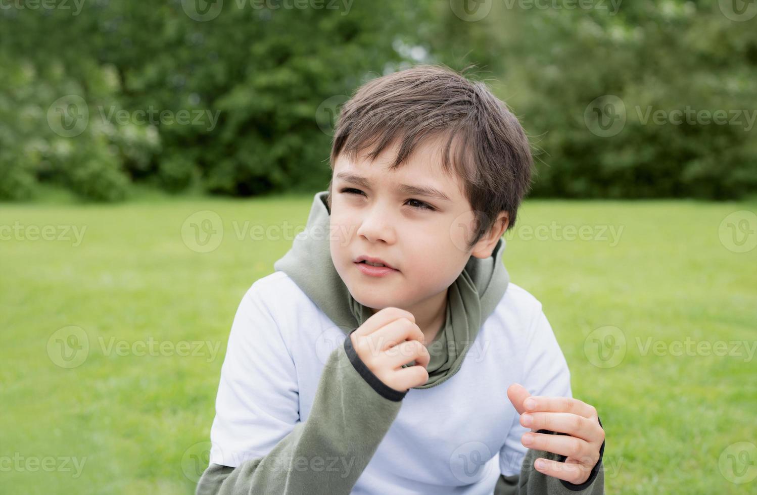 Portrait Young boy looking out deep in thought, School kid sitting  alone on green grass in garden, Cute Child playing outdoor with blurry green nature in the park photo