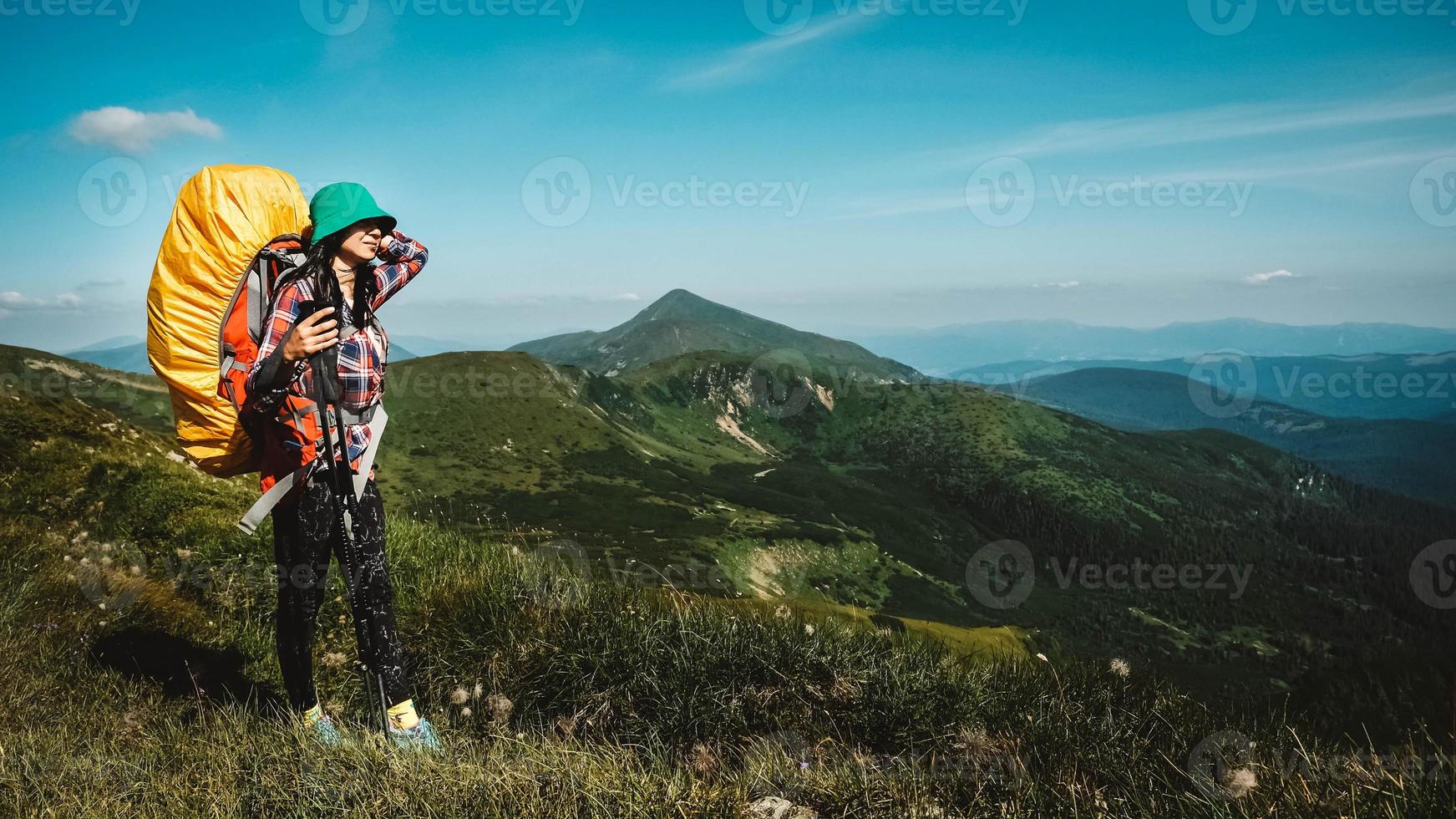 Woman tourist is walking on a hiking trail with a backpack against background of green mountains and sky photo