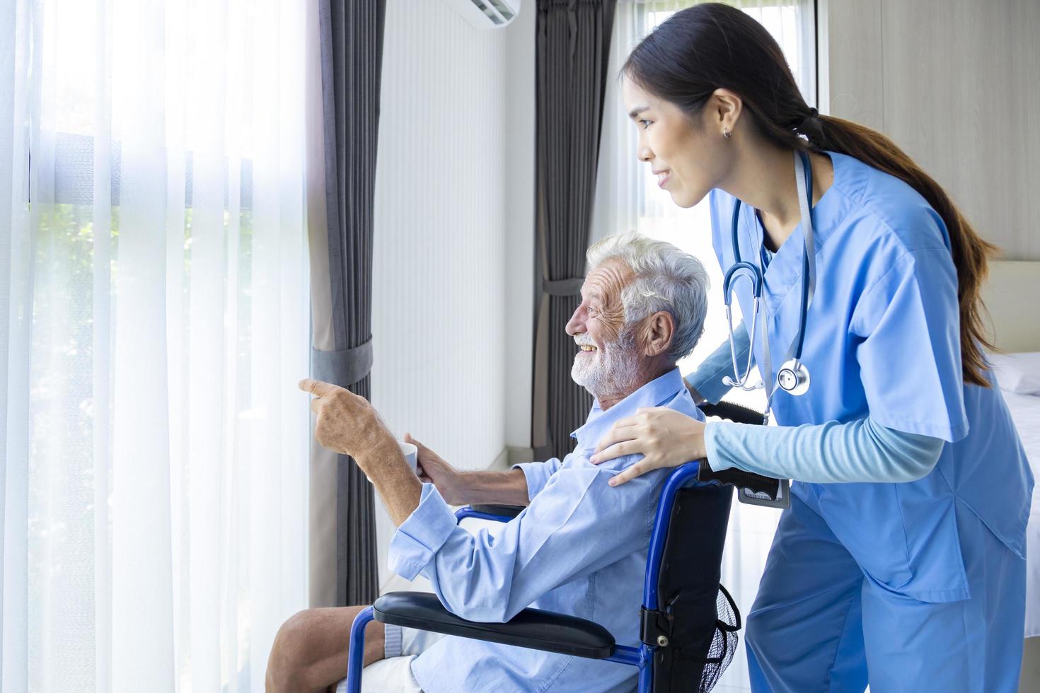 Hospice nurse is supporting Caucasian man in wheelchair while looking out the window in pension retirement center for home care rehabilitation and post treatment recovery process photo