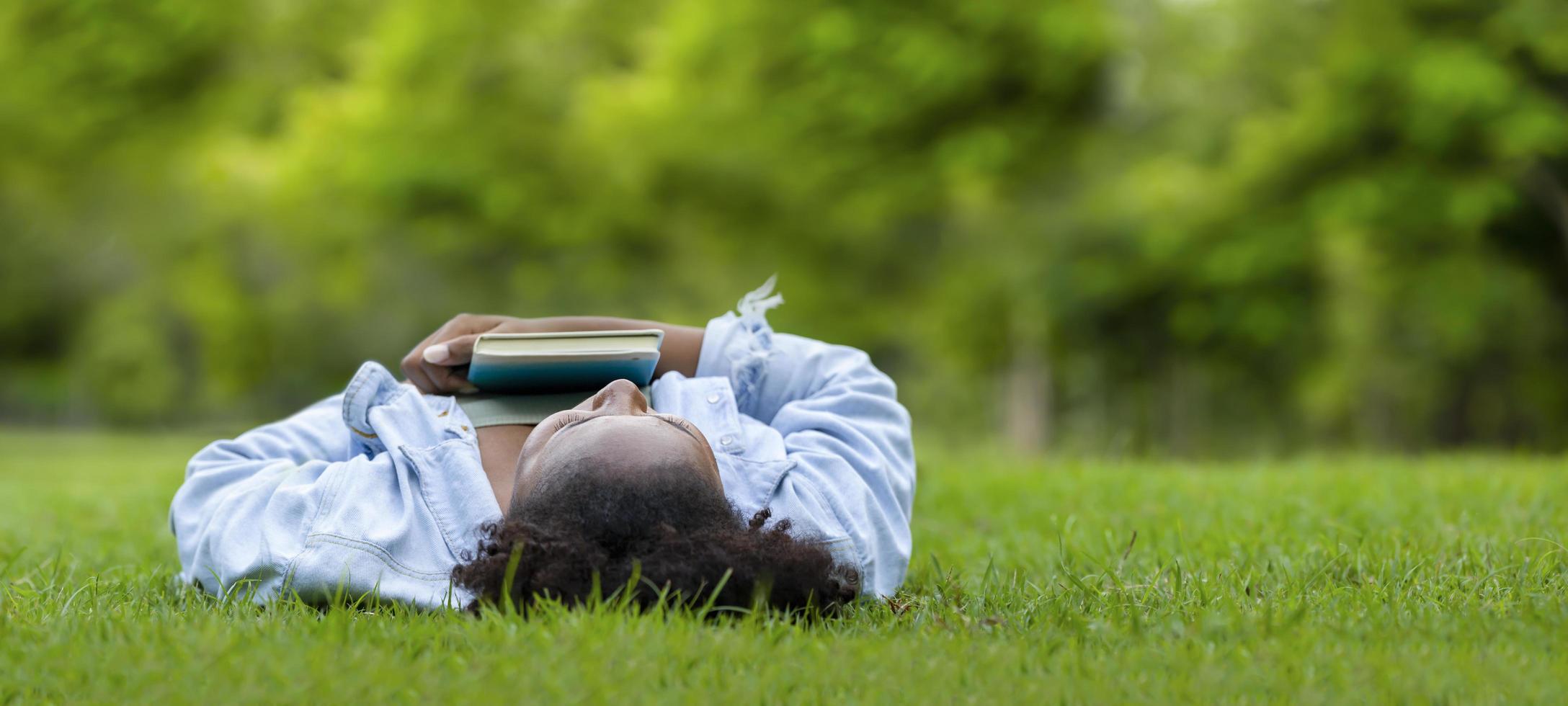African American woman is lying down in the grass lawn inside the public park holding book in her hand during summer for reading and education concept with copy space photo