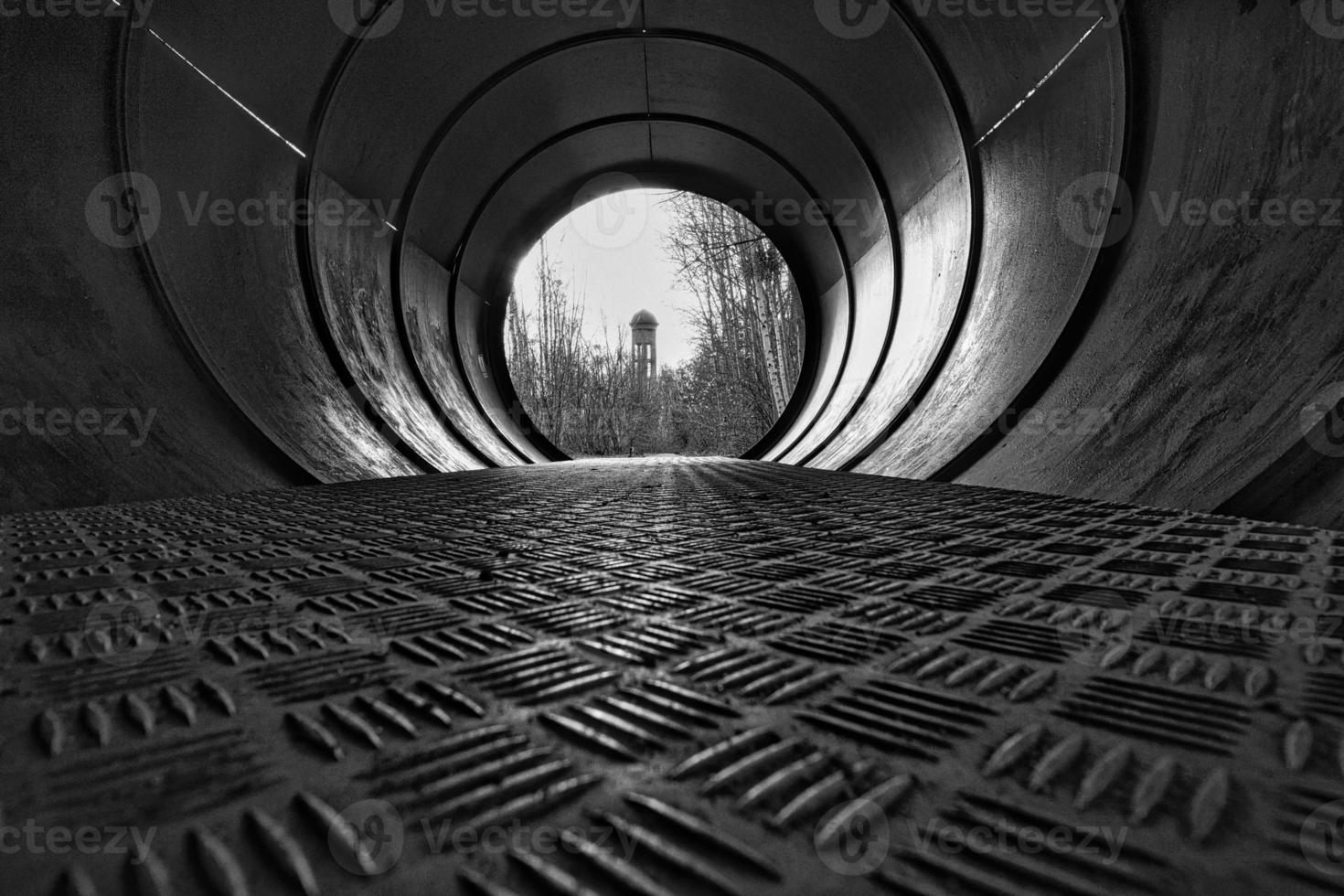 View through a metal pipe. In the background between trees is a water tower. photo