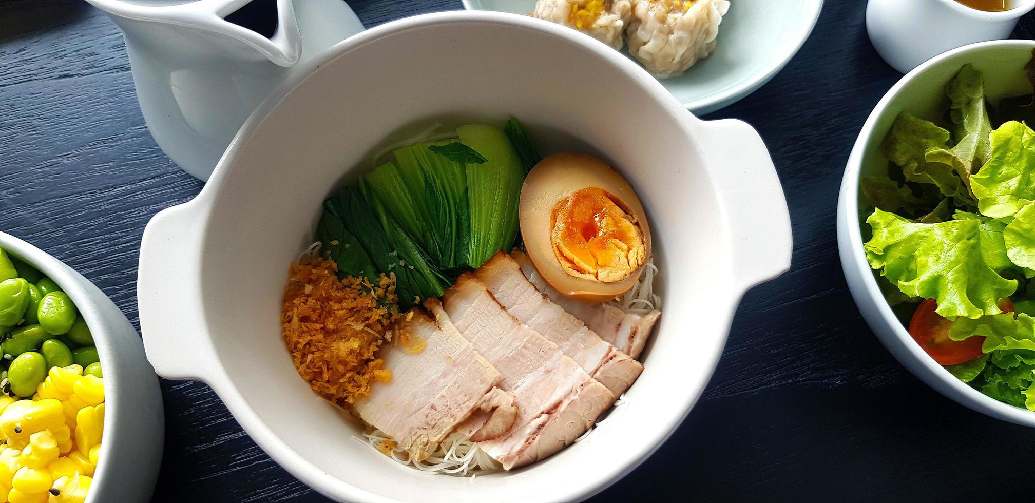 Top view of dry noodle with boiled belly pork, egg and garlic on white bowl with fresh salad, green cabbage, corn and bean. Flat lay of Breakfast set on black table background. Healty food concept. photo