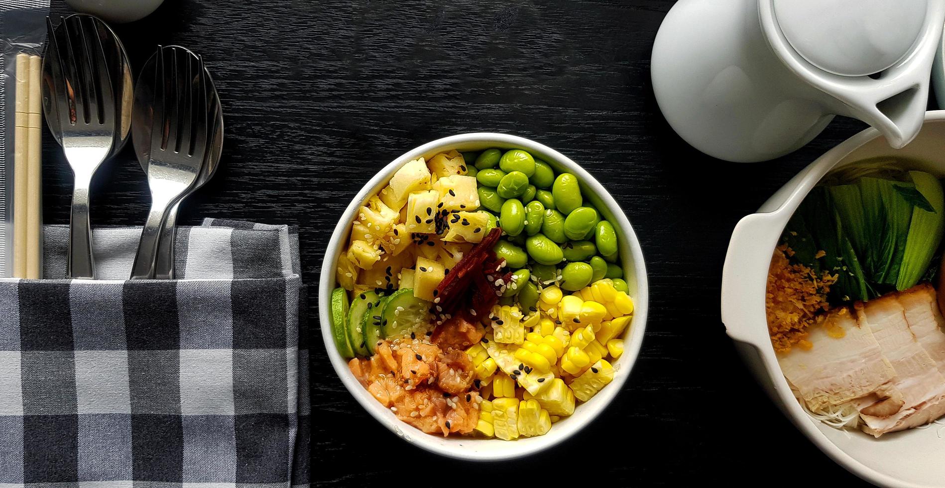 Flat lay of fresh salad with sliced of corn, salmon, green nut or bean,  pineapple, bacon, cucumber and topping with white and black sesame. Set of Healthy food  with spoon and fork put in napkin. photo