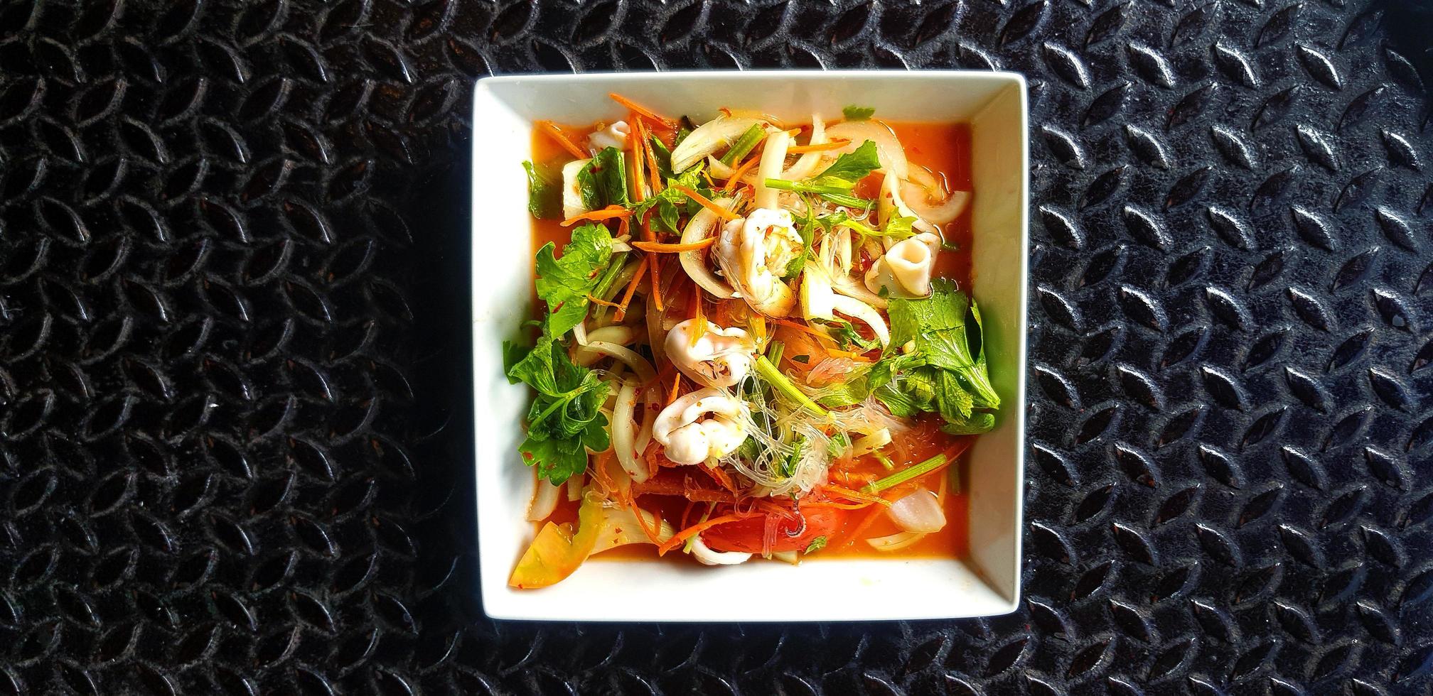 Ensalada tailandesa de mariscos picantes con fideos de vidrio, calamares, camarones, rodajas de tomate, cebolla, zanahoria y apio en un plato o plato blanco. comida asiática y famosa sobre fondo o papel tapiz de acero inoxidable negro. foto