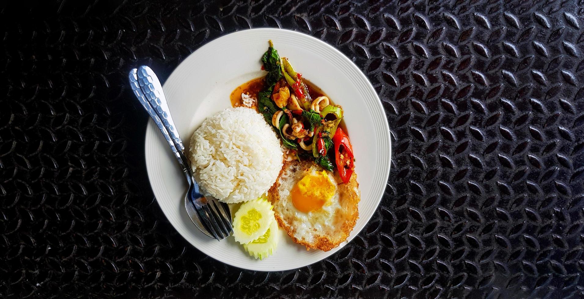 Top view of Stir Fried Basil with squid, rice, sliced cucumber and fried egg in Thai style on white plate with copy space. Flat lay of Famous and spicy food on black stainless steel background. photo