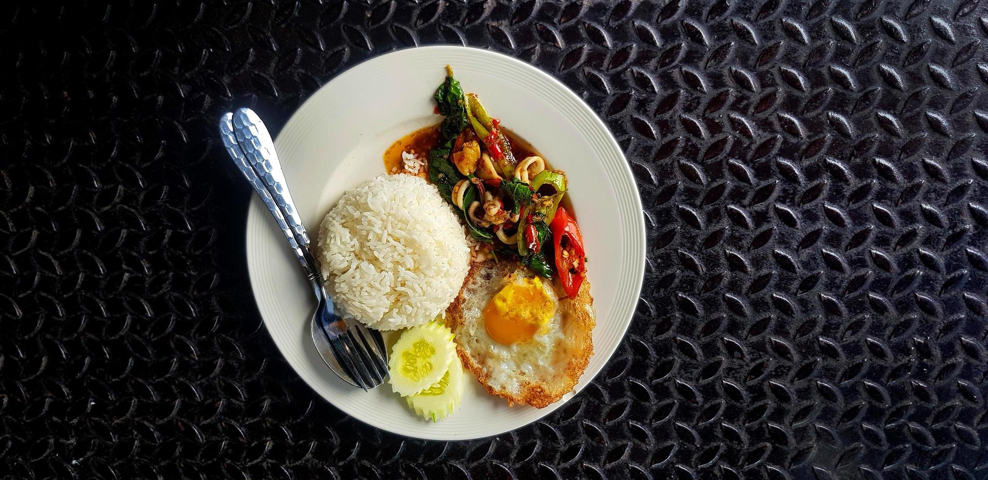 vista superior de albahaca salteada con calamares, arroz, pepino en rodajas y huevo frito al estilo tailandés en un plato blanco con espacio para copiar. endecha plana de comida famosa y picante sobre fondo negro de acero inoxidable. foto