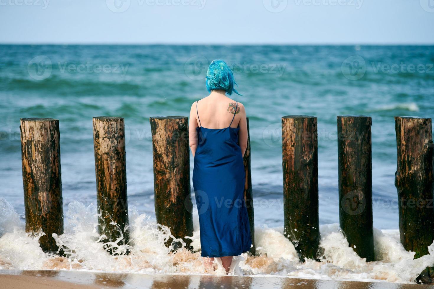 Young blue-haired woman in long dark blue dress standing on sandy beach looking at ocean horizon photo