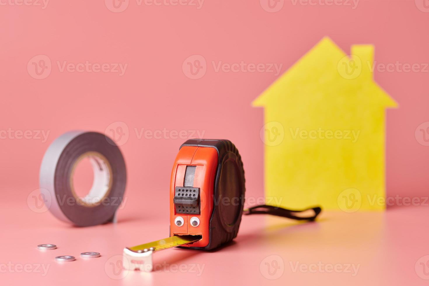 House renovation funny concept. Metal tape measure and other repair items. Home repair and redecorated concept. Yellow house shaped figure on pink background. photo