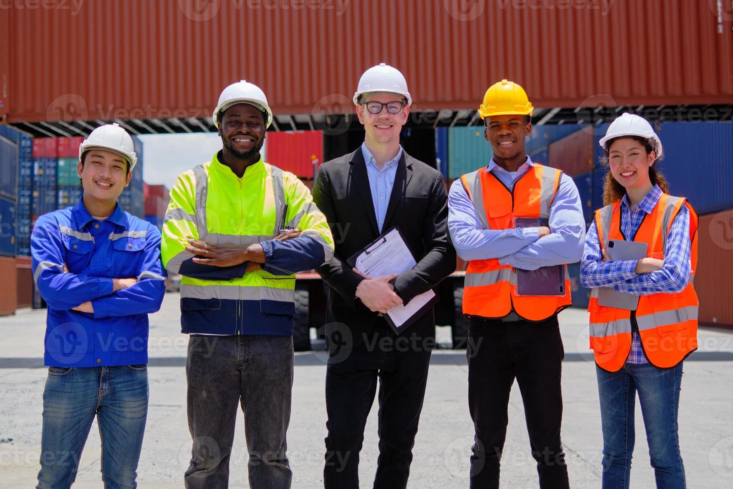 Group of multiracial workers team in safety uniforms, arms crossed and looking at camera at logistics terminal with many stacks of containers, loading shipping goods, cargo transportation industry. photo