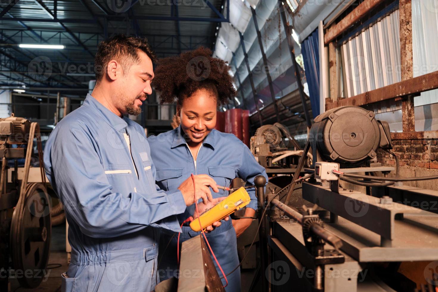 Asian male and female African American engineers in safety uniform work by inspecting machines' voltage current, checking, and maintaining at manufacture factory, electric system service occupations. photo