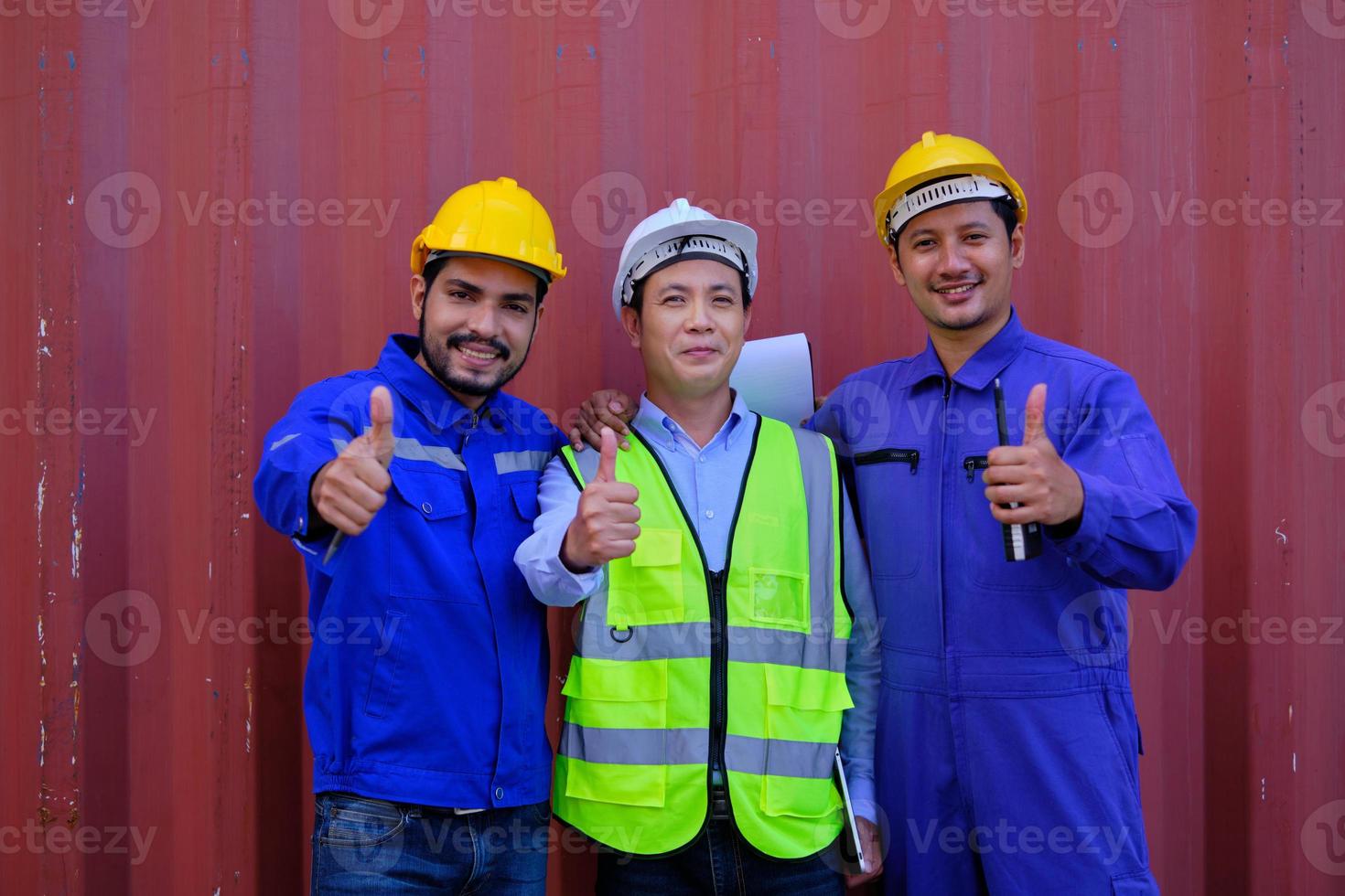 Portrait of three Asian industry workers with safety uniform and hard hats on metal sheet background, thumb up, look at camera and smile, cargo shipping, professional freight transportation business. photo
