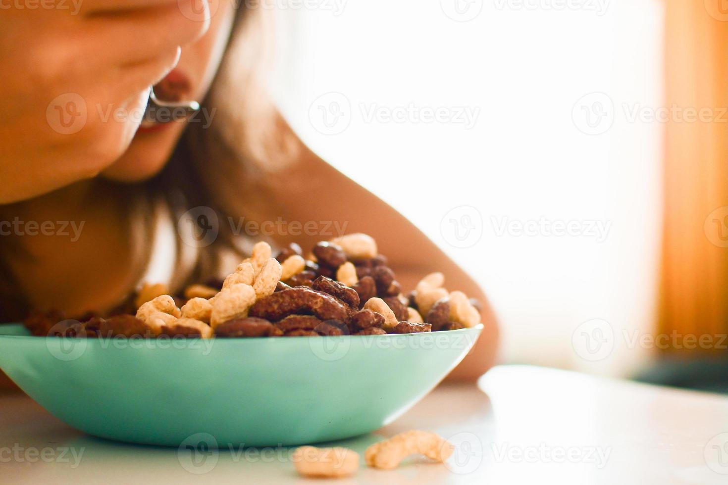 Close up view caucasian boy sit to eat cereals in kitchen on white table. Food in focus blurry people intentional bright background photo