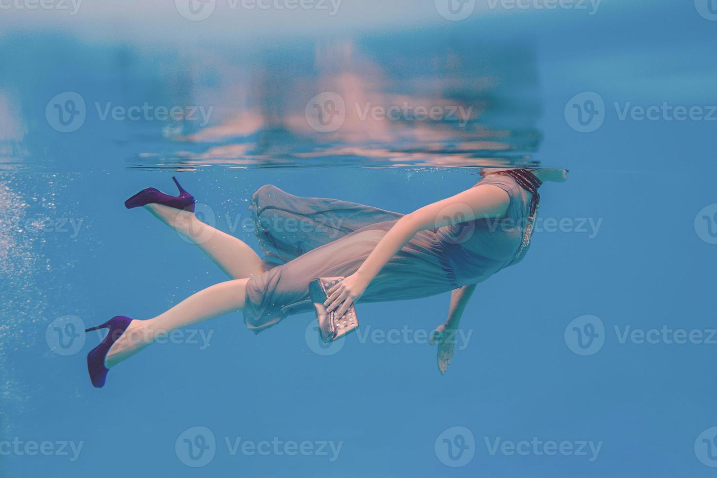 retrato artístico surrealista de una joven vestida de gris, bufanda con cuentas, bolso pequeño, tacones altos violetas bajo el agua en la piscina foto