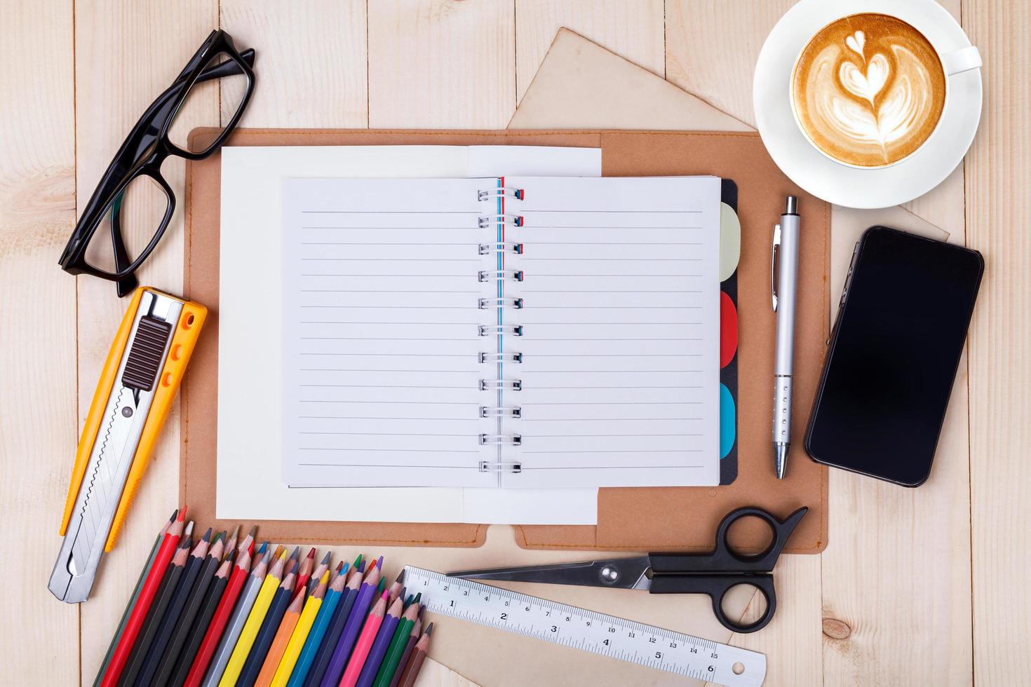 An open blank notebook with colour pencils, pen,smartphone and cup of coffee on wooden table photo