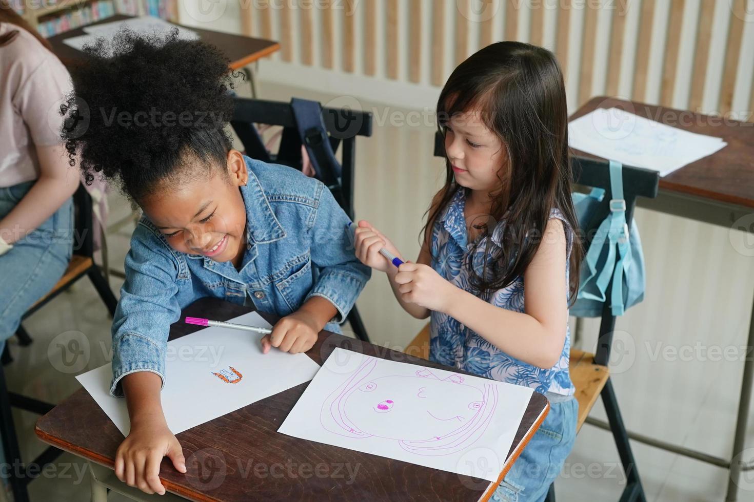 Preschool Girl Kid Drawing With Color Pencil On White Paper On Table In Classroom With Friends photo