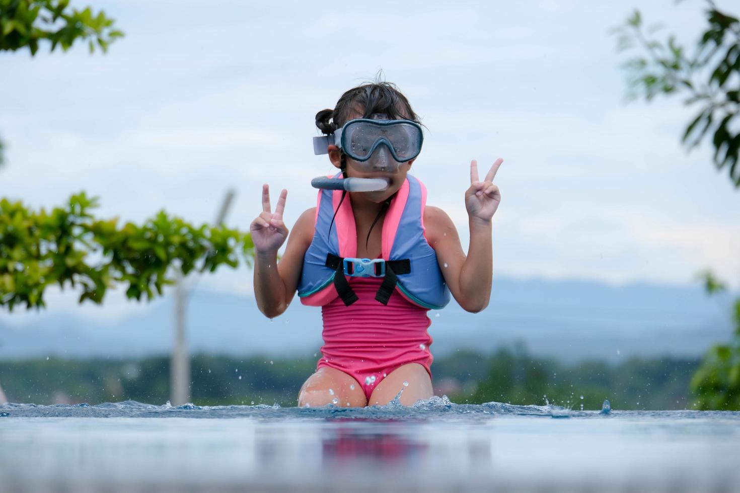Cute little girl wearing goggles is sitting by the pool preparing for a diving lesson. Happy little girl is swimming and playing in outdoor swimming pool. Healthy Summer Activities for Kids. photo