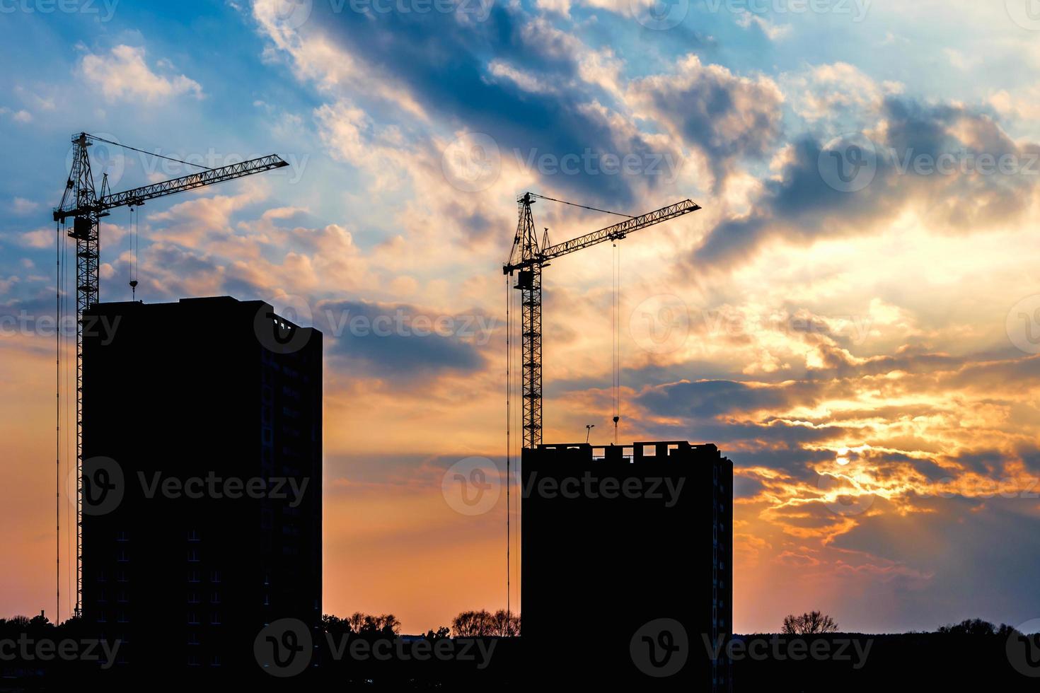 silhouette of tower cranes and unfinished multi-storey high buildings under construction site in the rays of the setting sun of beautiful sunset photo