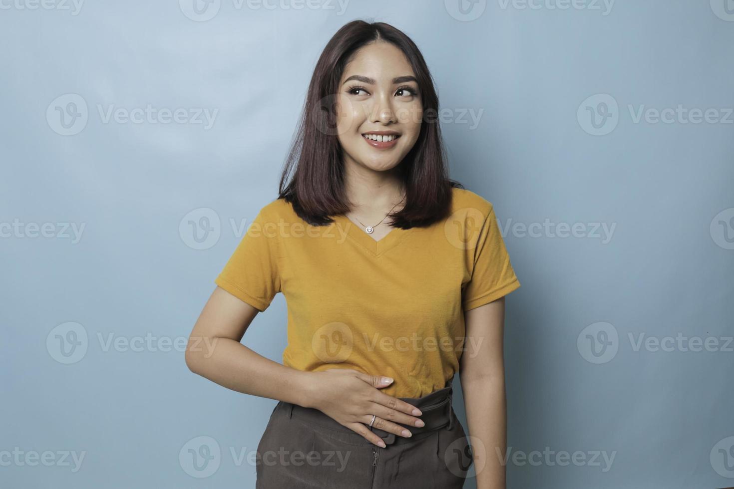 Pleased cheerful Asian woman keeps hand on belly feels full after delicious dinner dressed casually stands thoughtful against blue background. photo