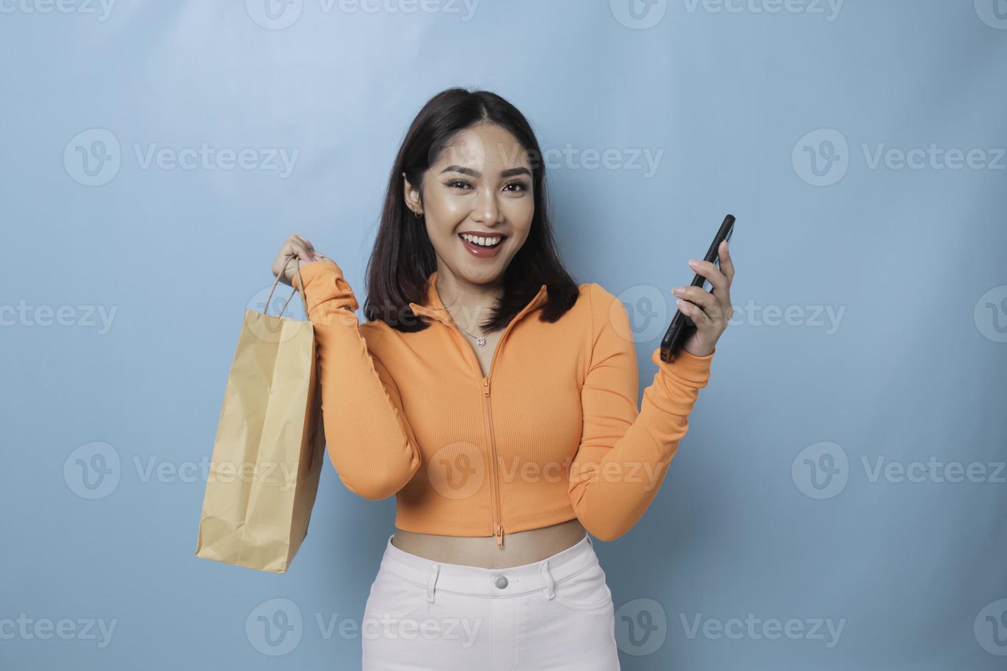 Portrait Asian happy beautiful young woman standing excited holding online shopping bag and smartphone on hand, studio shot isolated on blue background photo